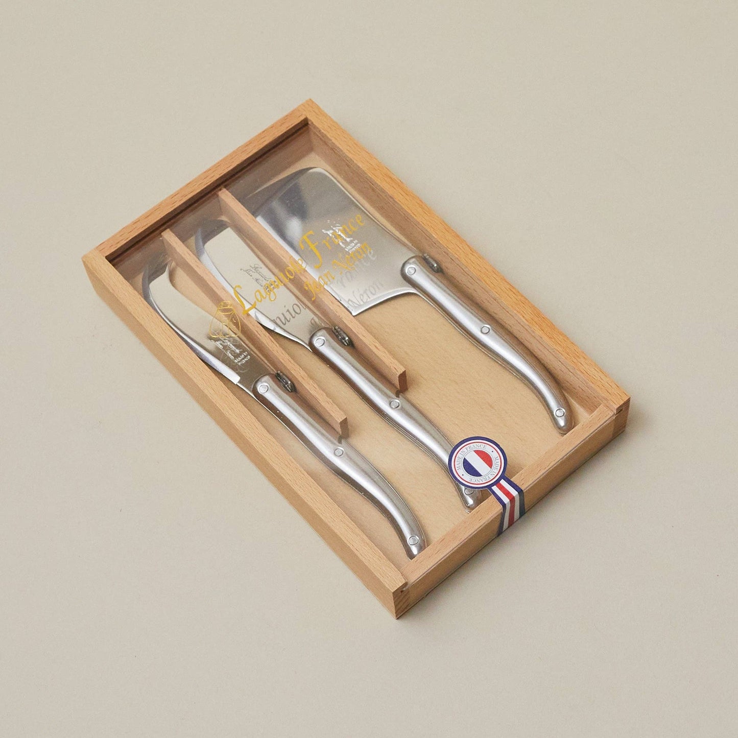 Set of four silver cheese knives in a wooden box with a brand logo on a beige background
