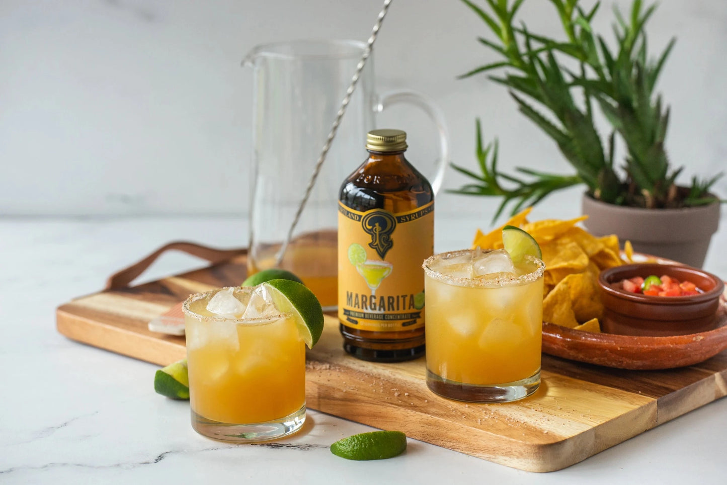 Two margaritas on a wooden cutting board with a bottle of margarita mix and snacks.