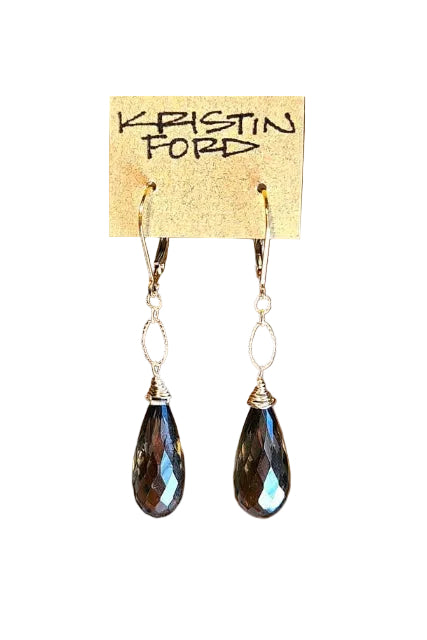 A pair of dangling earrings with faceted dark teardrop-shaped beads and silver-toned hooks and wire details, hanging from a tan display card labeled KRISTIN FORD against a warm brown blurred background.
