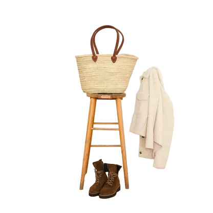 Woven basket on a wooden stool with brown boots and a beige coat against a white wall.