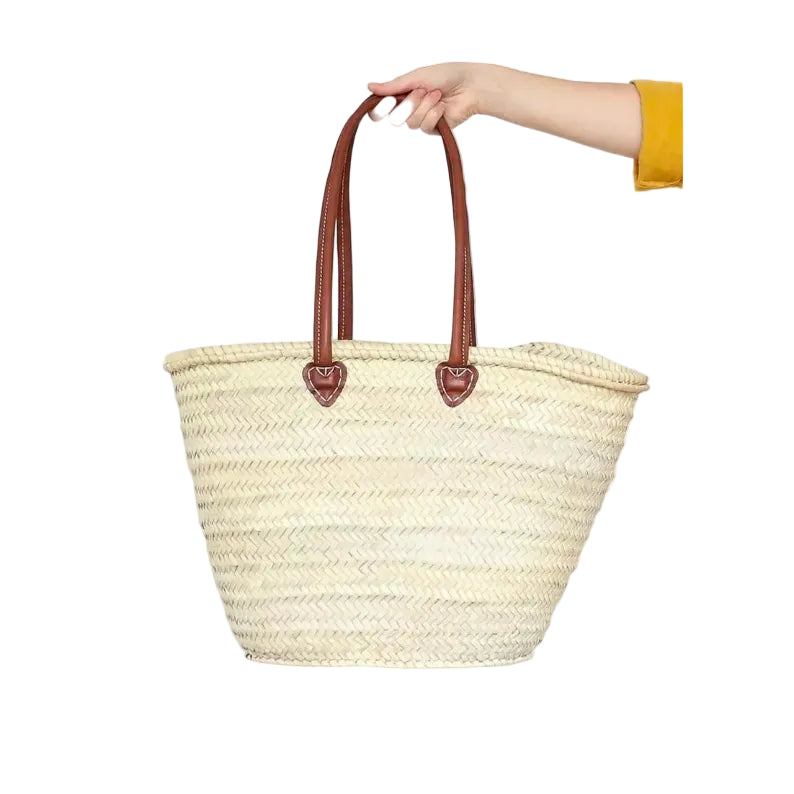 Woven straw tote bag with brown handles held by a hand on a white background
