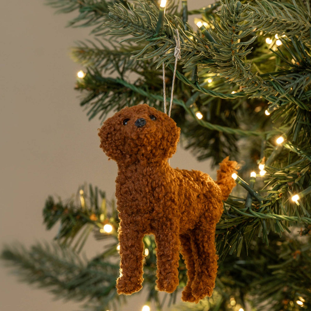 A brown curly-fur dog Christmas ornament hanging from a green Christmas tree with warm white string lights against a soft beige background.
