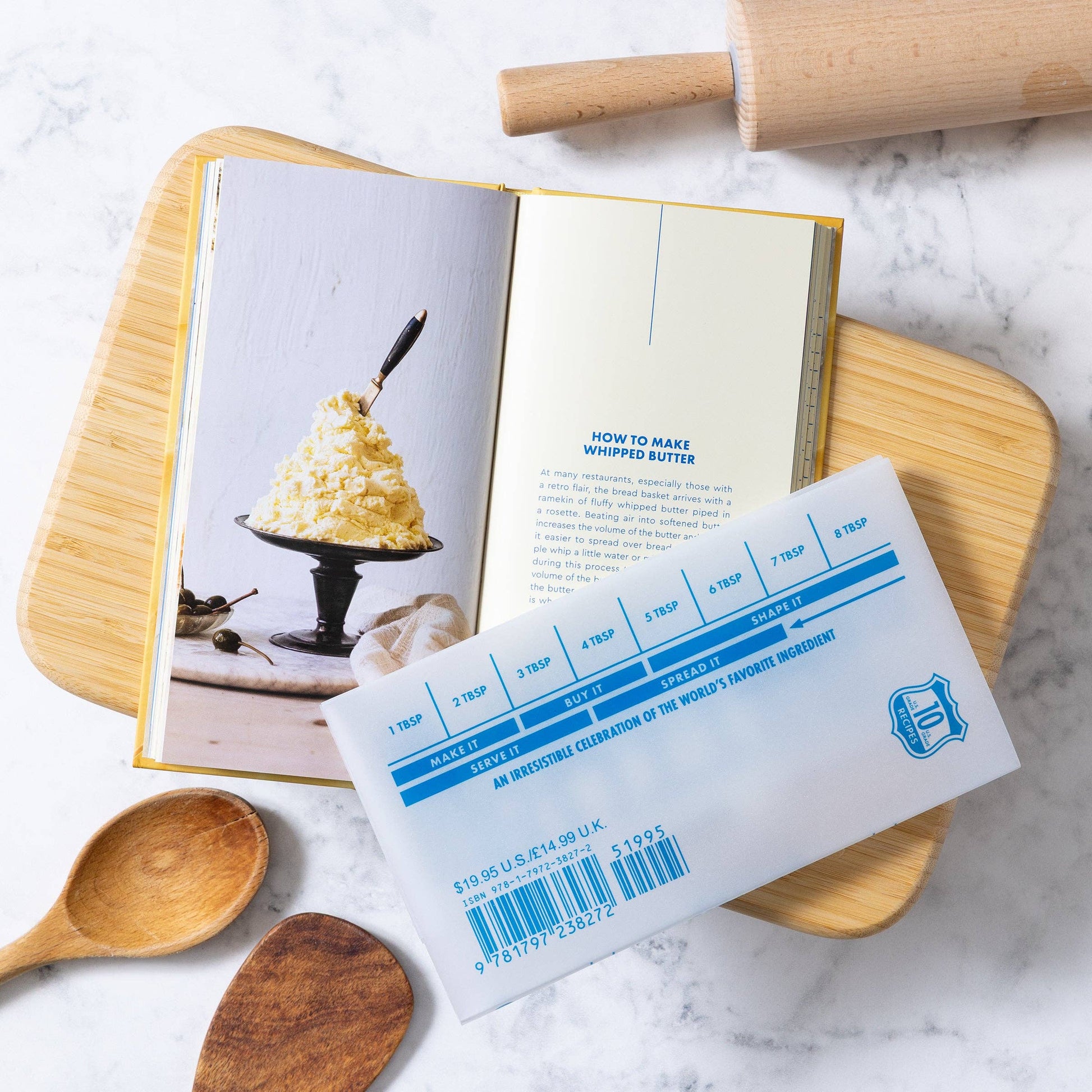Butter container with measurement scale on a cookbook open to a recipe for whipped butter, surrounded by wooden spoons and a rolling pin.