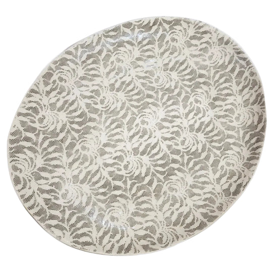 An oval ceramic plate featuring an intricate, repeating floral lace-like pattern in cream and light gray.