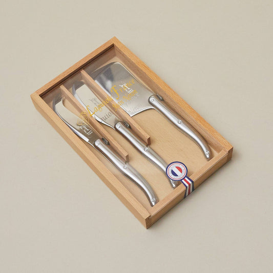 Set of four silver cheese knives in a wooden box with a brand logo on a beige background