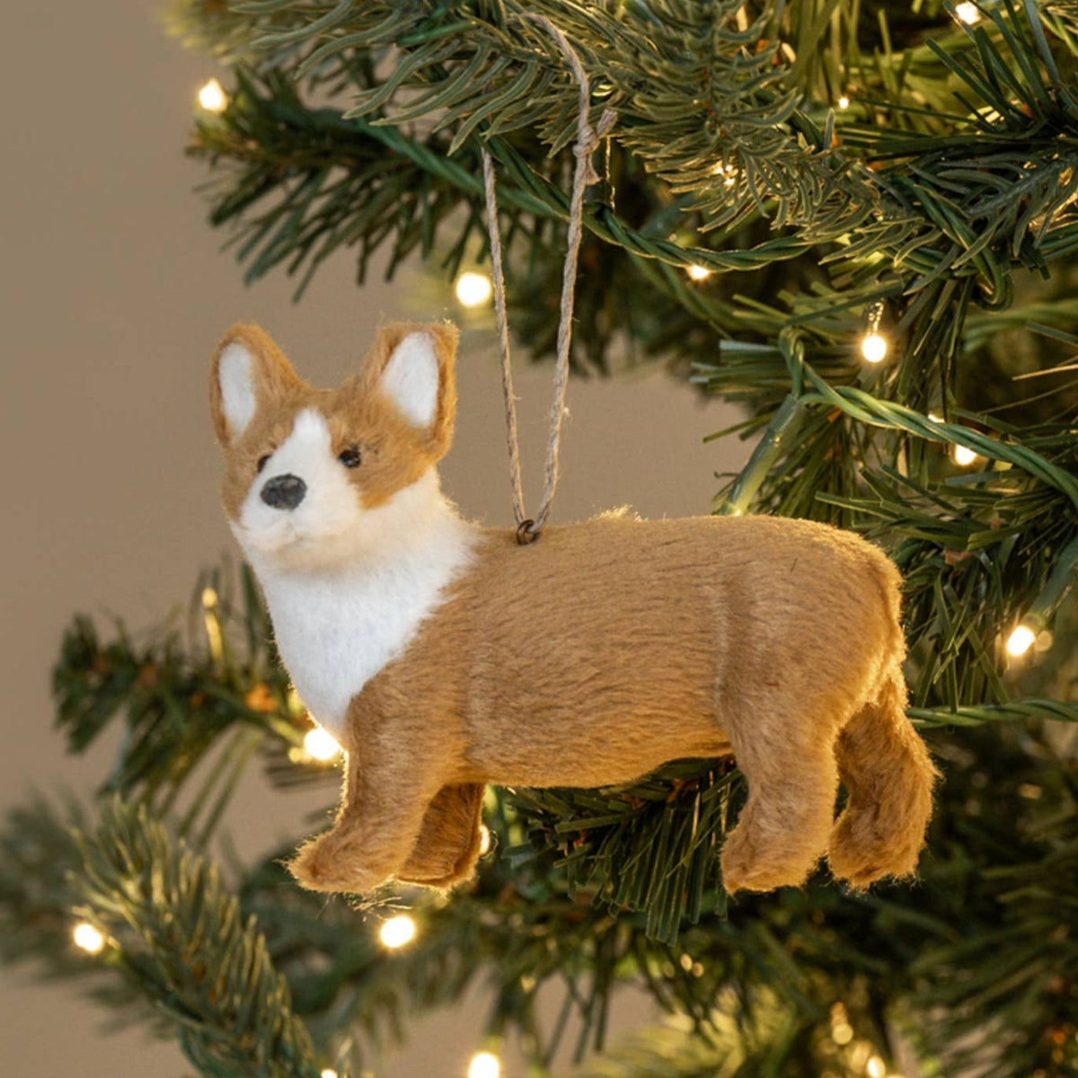 A plush Corgi Christmas ornament hanging from a string on a decorated Christmas tree with warm white lights.
