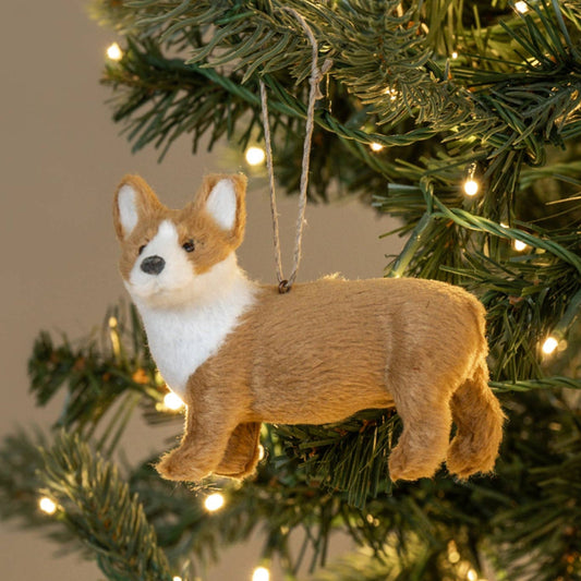 A plush Corgi Christmas ornament hanging from a string on a decorated Christmas tree with warm white lights.