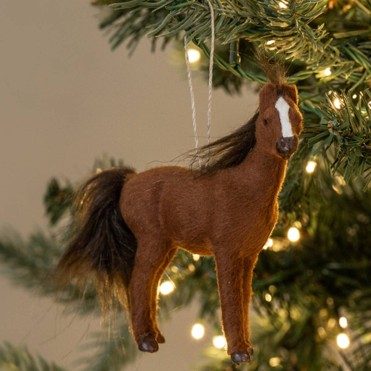 A brown horse-shaped Christmas ornament with a white blaze hangs from a lit green Christmas tree against a warm beige background.