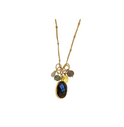 A gold charm necklace featuring a large oval black labradorite stone with blue iridescence, accompanied by smaller raw gemstone charms and a gold coin charm, displayed on a light beige background.