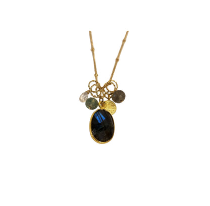 A gold chain necklace with a cluster of charms, featuring a large oval black labradorite stone pendant, a small gold coin charm, a clear crystal bead, a green stone bead, and a dark gray stone bead, displayed on a light beige background.