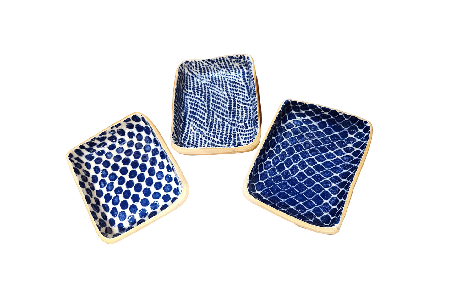 Three square ceramic plates with rounded corners in blue and white patterns, arranged on a rustic wooden table. One plate features a blue polka dot pattern, while the other two feature blue and white wavy and lattice-like patterns.