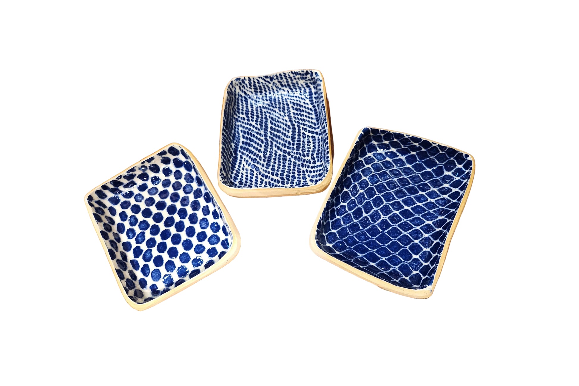 Three square ceramic plates with rounded corners in blue and white patterns, arranged on a rustic wooden table. One plate features a blue polka dot pattern, while the other two feature blue and white wavy and lattice-like patterns.