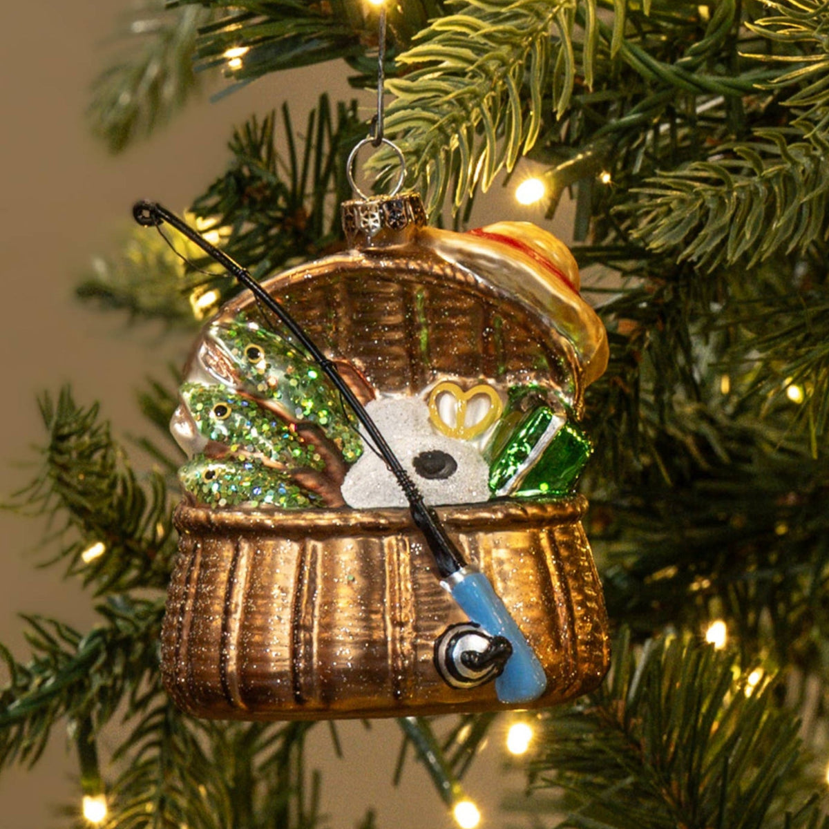 A Christmas ornament shaped like a small wicker basket containing a white cat wearing a green hat and blue scarf, hanging on a lit green Christmas tree against a warm beige background.