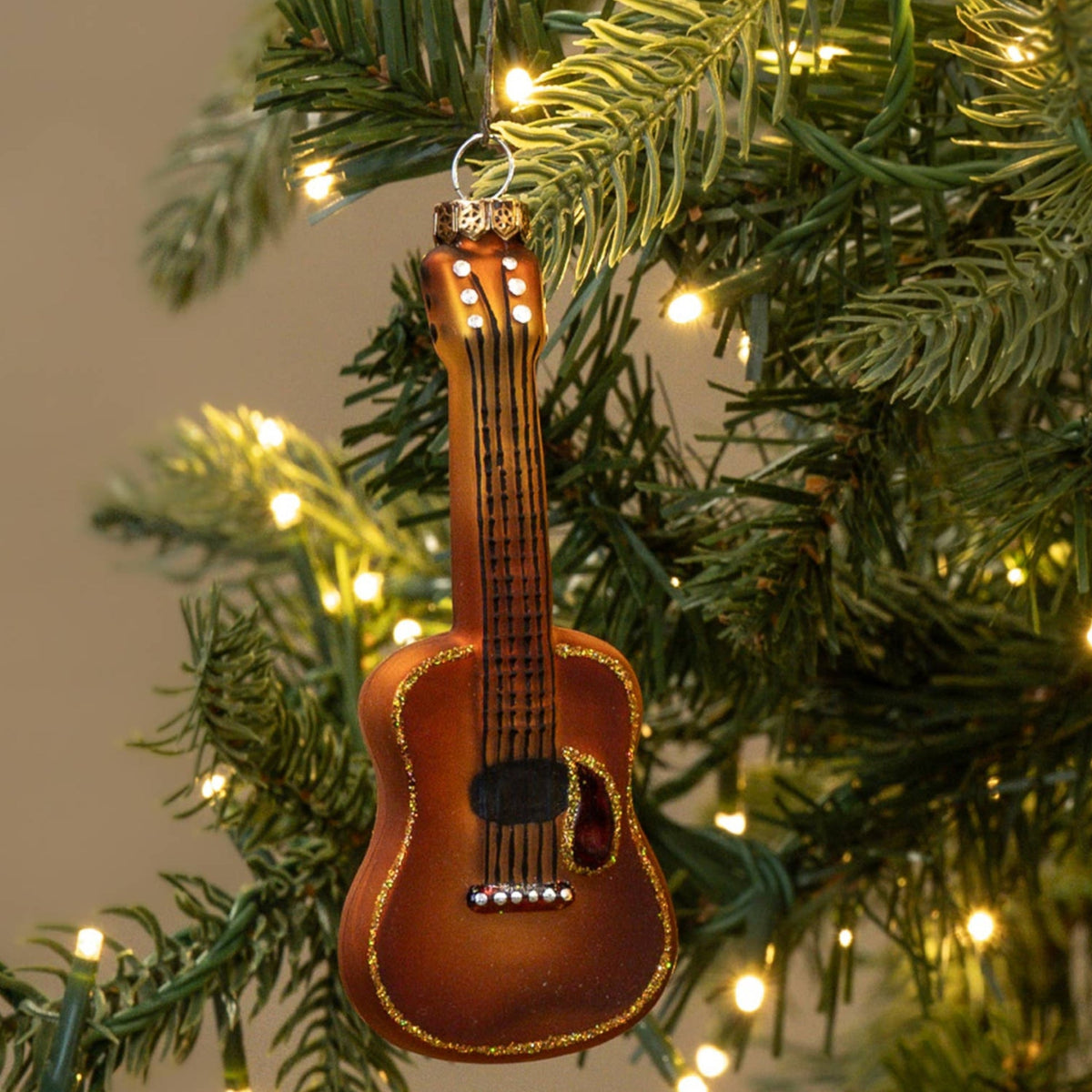 A small brown ukulele Christmas ornament hanging from the green branches of a lit Christmas tree.