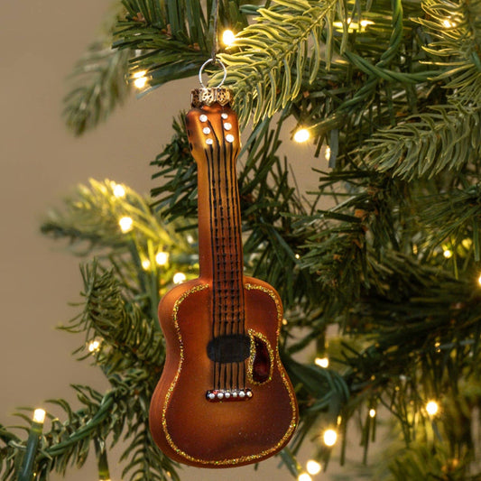 A small brown ukulele Christmas ornament hanging from the green branches of a lit Christmas tree.