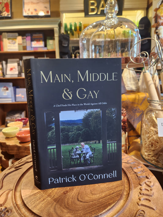 The book 'Main, Middle & Gay' by Patrick O'Connell stands on a carved wooden tray in a cozy, rustic shop setting. The book cover is dark navy blue with white text and a central photograph of a couple sitting by a window.