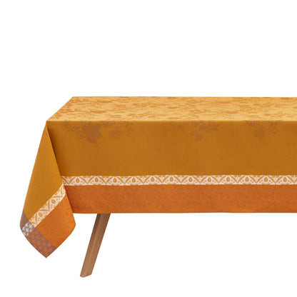 A mustard yellow tablecloth with a subtle damask pattern and a white geometric border, draped over a table with a wooden leg visible against a white background.
