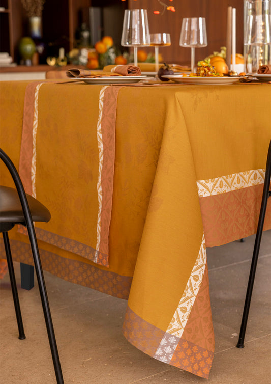 A mustard yellow tablecloth with orange and white patterned borders, draped over a dining table set with wine glasses, plates, and autumn decorations.