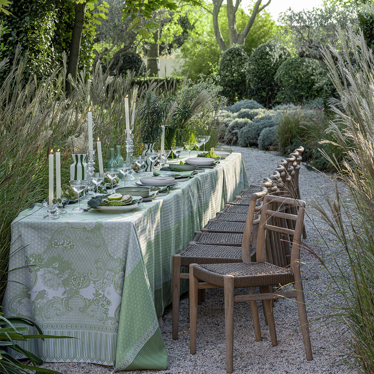 Dining table set for a meal outdoors with green tablecloth and chairs in a garden setting.