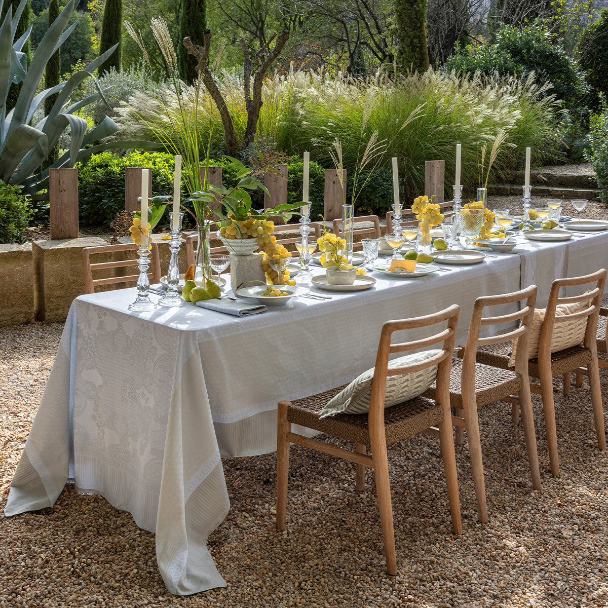 Dining table set for a meal outdoors with white coated tablecloth, yellow flowers, and candles in a garden setting.