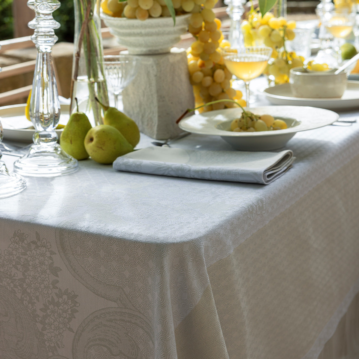 Elegant table setting with white tablecloth, fruit, and glasses on a patterned surface.