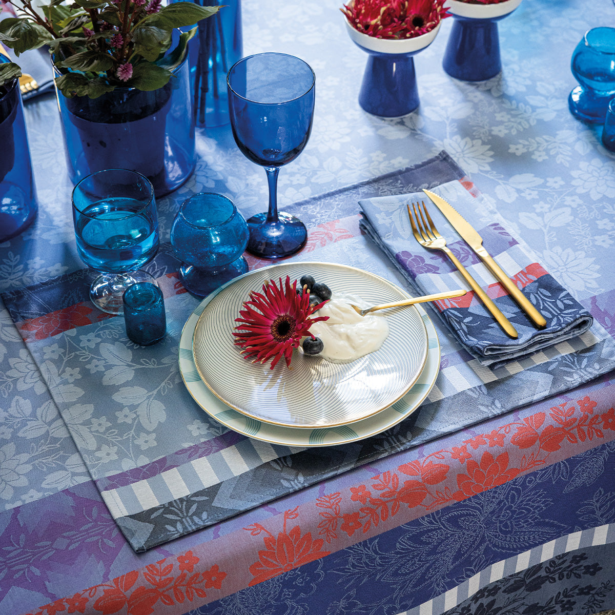 Decorative table setting with blue glasses, floral plates, and colorful coated placemat and coated tablecloth.
