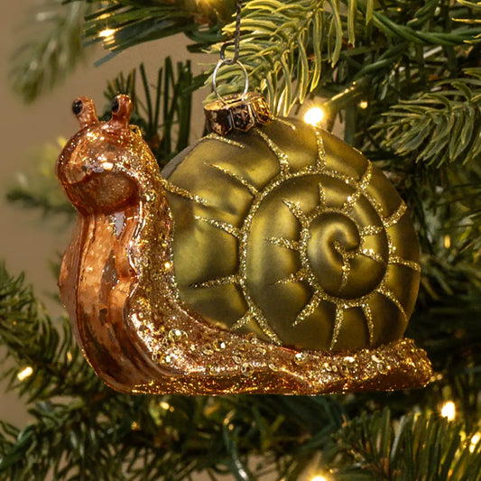 A snail-shaped Christmas ornament with a metallic gold shell and glittery coppery-brown body, hanging on a green Christmas tree with warm string lights.