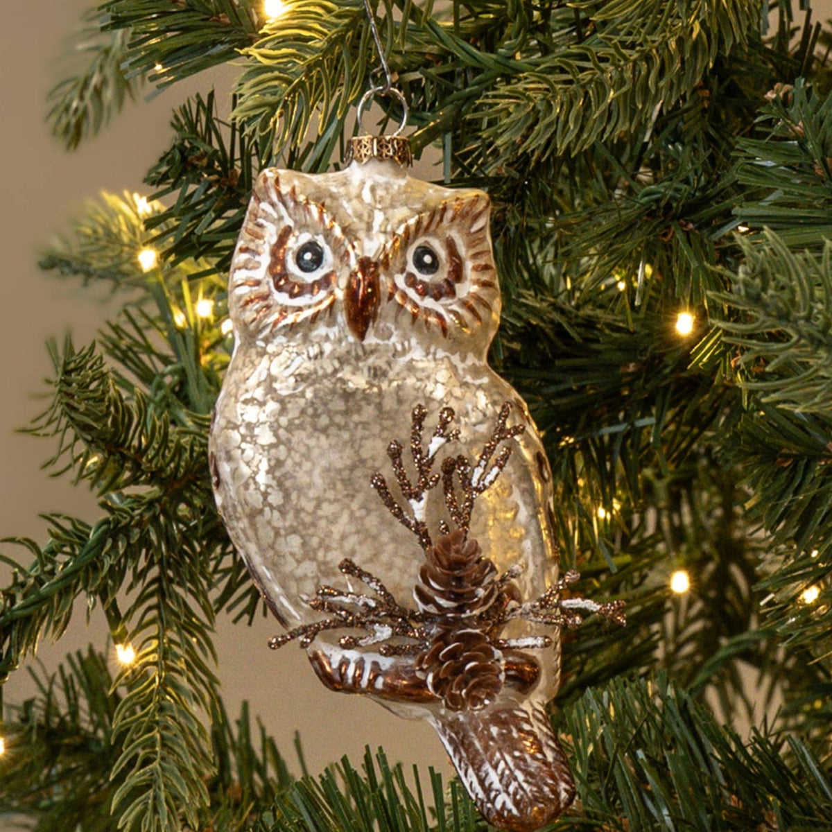 A glittery owl Christmas ornament hanging on a green artificial Christmas tree with warm white string lights against a plain beige background.