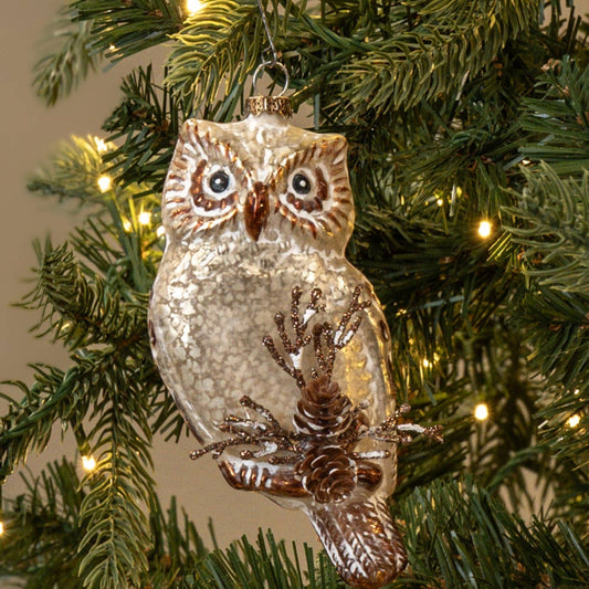 A glittery owl Christmas ornament hanging on a green artificial Christmas tree with warm white string lights against a plain beige background.
